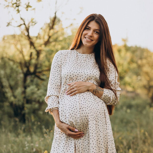 Femme enceinte souriante en robe blanche à pois tenant son ventre arrondi dans la nature